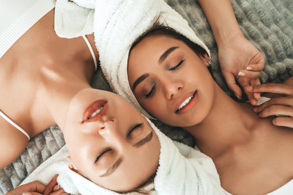 Two young beautiful smiling women in white bathrobes and towels on head.Sexy carefree models lying on bed in posh apartment. They doing beauty treatments at home in towels on heads.After face massage